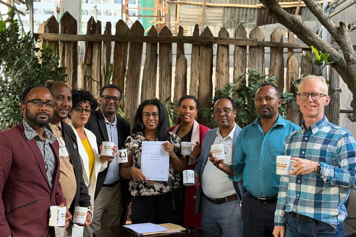A group from Mother to Mother standing together outdoors, holding branded mugs and a signed document.